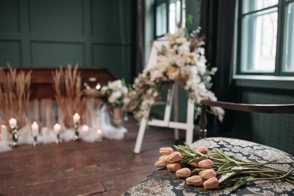 A soft-focus floral arrangement scene featuring a bouquet of pale pink tulips with green stems resting on an ornate, dark-patterned upholstered chair in the foreground. In the background, a table deco
