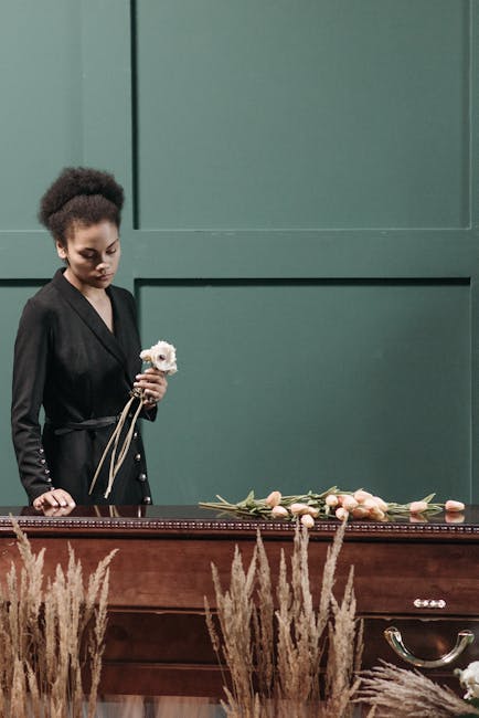 A young woman with curly dark hair, dressed in a black coat, is holding a small white flower bouquet with a ribbon, standing beside a wooden casket. On top of the casket, there are several floral arra