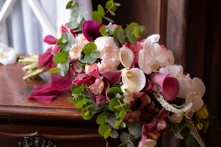 A floral arrangement featuring pink and white flowers, including calla lilies, orchids, and carnations, accented with green eucalyptus leaves. The bouquet is resting on a polished wooden surface, with