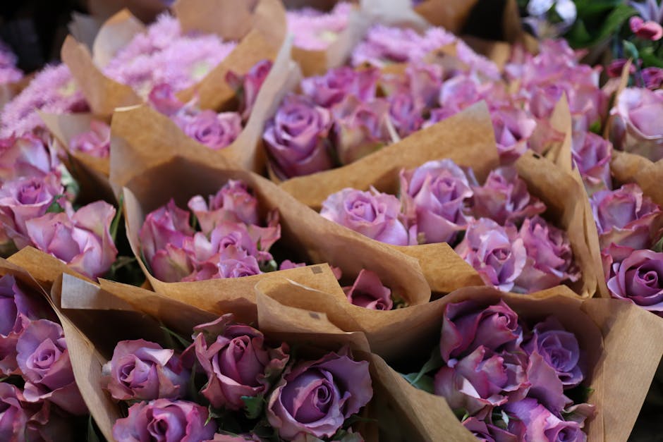 A collection of fresh pink and lavender roses arranged in small, open brown paper wraps, ready for floral gift delivery. The roses have tightly packed petals with a velvety texture, showing shades of 