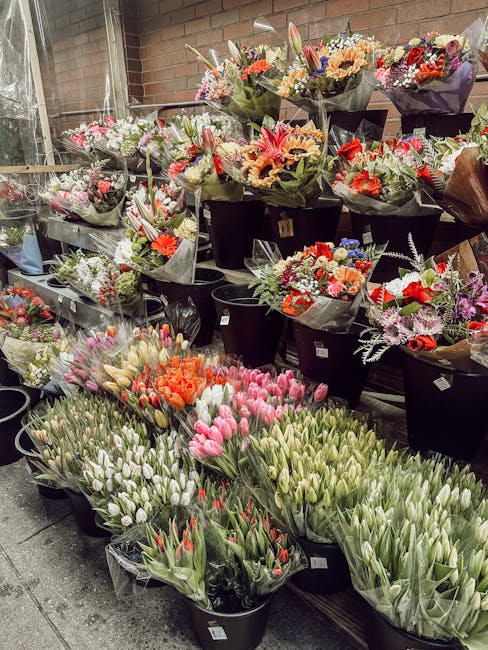 An extensive display of fresh flowers arranged in black buckets and wrapped bouquets inside a floral shop, with a variety of colorful blooms including pink and white tulips, orange and pink lilies, pu