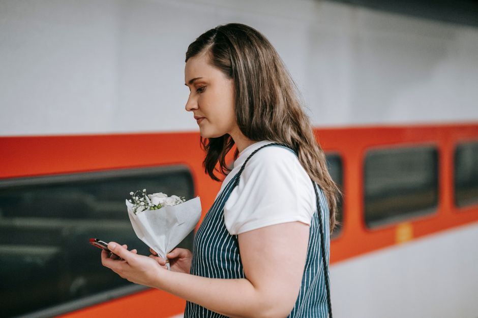 A young woman with long brown hair, dressed in a white T-shirt and a blue and white striped apron, is holding a small bouquet of fresh white flowers wrapped in white paper with green foliage accents. 
