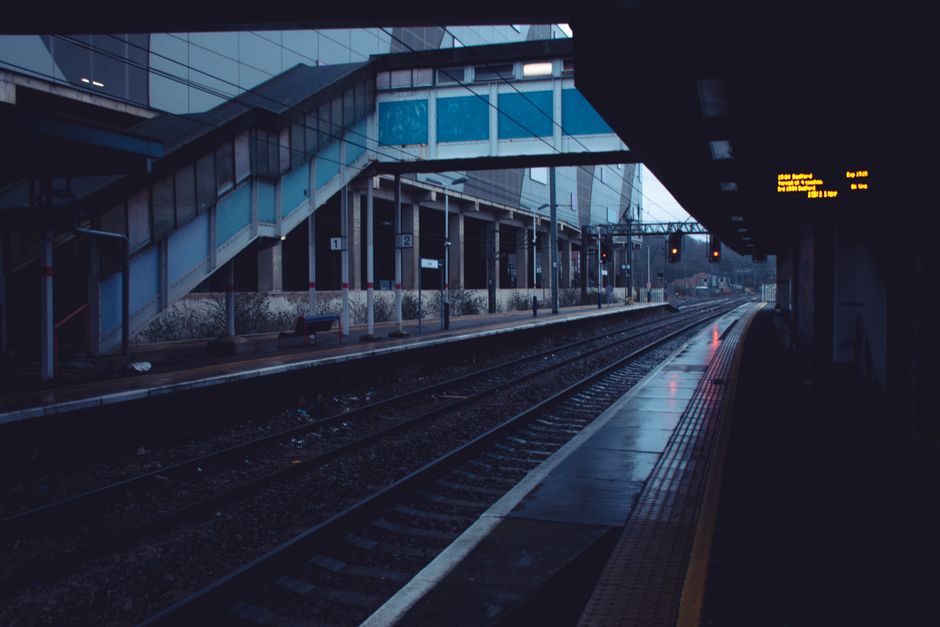 An empty train station platform near Luton Train Station LU2 during early morning or late evening, with wet pavement reflecting dim lighting. The station features modern glass and metal structures, in